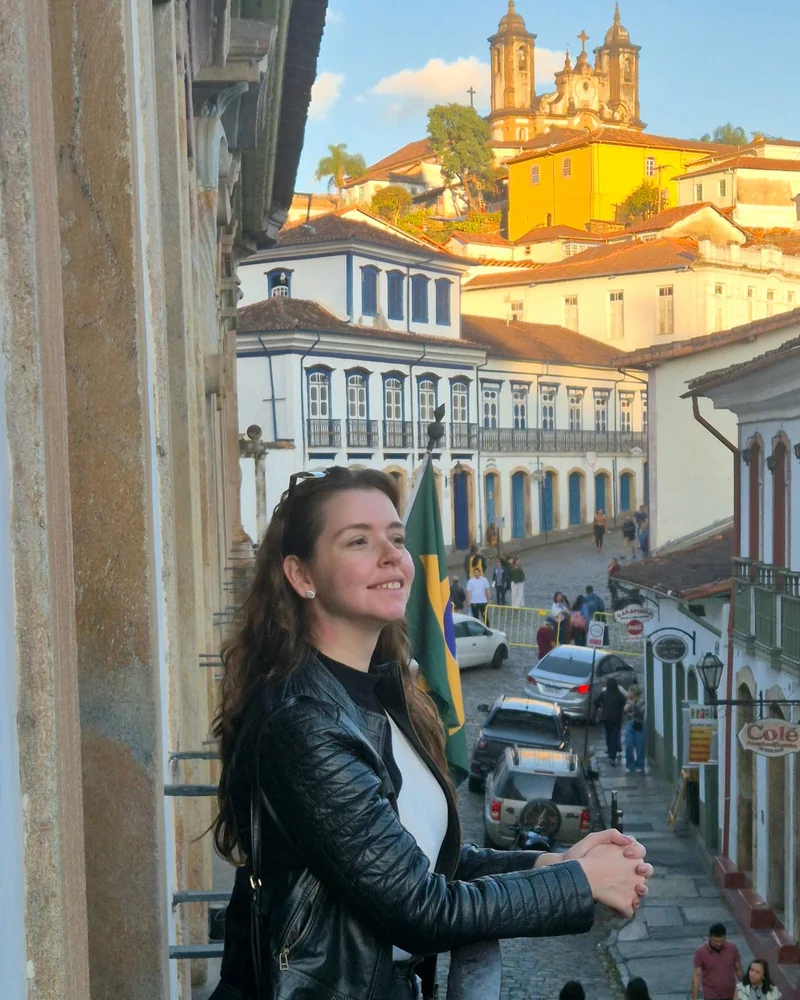 Júlia Salles sorrindo numa sacada, com casario colonial e uma igreja ao fundo